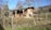 A rustic barn surrounded by a wire fence with several goats standing near the barn and chickens scattered across the grassy area. The background features leafless trees and a gently sloping hill under a clear blue sky.