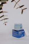 A skincare product displayed on a neutral background. The product consists of a jar with a translucent lid, sitting on a blue box featuring text and branding details. Nearby, some out-of-focus green leaves are visible, adding a natural element to the composition.