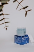 A skincare product displayed on a neutral background. The product consists of a jar with a translucent lid, sitting on a blue box featuring text and branding details. Nearby, some out-of-focus green leaves are visible, adding a natural element to the composition.