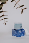 A skincare product displayed on a neutral background. The product consists of a jar with a translucent lid, sitting on a blue box featuring text and branding details. Nearby, some out-of-focus green leaves are visible, adding a natural element to the composition.