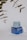 A skincare product displayed on a neutral background. The product consists of a jar with a translucent lid, sitting on a blue box featuring text and branding details. Nearby, some out-of-focus green leaves are visible, adding a natural element to the composition.