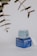 A skincare product displayed on a neutral background. The product consists of a jar with a translucent lid, sitting on a blue box featuring text and branding details. Nearby, some out-of-focus green leaves are visible, adding a natural element to the composition.
