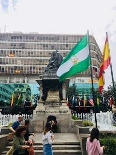 A large statue stands in front of a modern building adorned with multiple flags, including prominent green and white and red and yellow ones. A group of people walks in front of a series of fountains, adding a sense of motion to the scene.