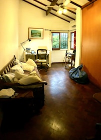 A cozy room with a wooden floor and high ceiling. There is a small wooden bench with cushions on the left side, and a desk with a lamp, books, and a laptop near the window. A modern office chair is positioned at the desk. The window is flanked by white curtains and provides a view of greenery outside. There is a ceiling fan with lights hanging from the beams, and a bean bag sits on the right side near a wooden cabinet.