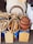 A collection of wooden kitchen utensils and cutting boards displayed in wicker baskets along with large clay pots and circular wooden boards. The items are arranged on top of large tins of cooking oil, next to stacks of folded towels. There is a rustic, organized feel to the setup.