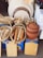 A collection of wooden kitchen utensils and cutting boards displayed in wicker baskets along with large clay pots and circular wooden boards. The items are arranged on top of large tins of cooking oil, next to stacks of folded towels. There is a rustic, organized feel to the setup.