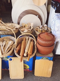 A collection of wooden kitchen utensils and cutting boards displayed in wicker baskets along with large clay pots and circular wooden boards. The items are arranged on top of large tins of cooking oil, next to stacks of folded towels. There is a rustic, organized feel to the setup.