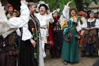 A group of people dressed in elaborate historical costumes stand together outdoors. Some hold musical instruments like a tambourine, while others are smiling and engaging with each other. A woman in the center holds a red and yellow rose. The crowd appears to be gathered in a festive, lively atmosphere under a tree.
