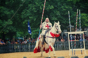 A knight in medieval armor rides a white horse in a jousting tournament. The knight wears a tunic with a red cross and holds a lance, surrounded by wooden structures and a crowd of onlookers. The event takes place outdoors, with green trees in the background.