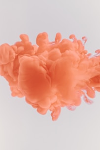Swirling orange ink is suspended in a light, possibly white, liquid, creating soft, cloud-like formations with smooth transitions between lighter and darker shades of orange.