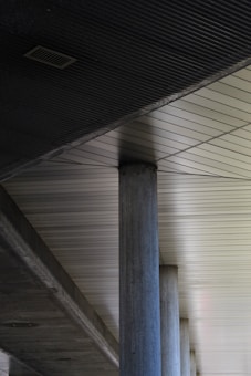 A series of industrial concrete columns supporting a modern ceiling with linear patterns and metallic tones. The ceiling features geometric lines that create a structured and minimalist aesthetic.