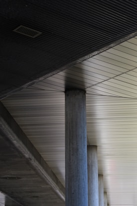 A series of industrial concrete columns supporting a modern ceiling with linear patterns and metallic tones. The ceiling features geometric lines that create a structured and minimalist aesthetic.