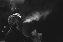 A person is exhaling smoke in a dimly lit environment, creating a dramatic effect with the interplay of light and shadows. The image is in black and white, emphasizing the contrast and mood.