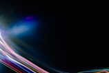 A dark canvas is intersected by vibrant streaks of colored light, including reds, blues, greens, and purples, creating a dynamic and futuristic atmosphere. The light patterns appear to move across the image, giving a sense of motion.