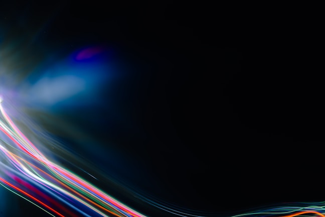 A dark canvas is intersected by vibrant streaks of colored light, including reds, blues, greens, and purples, creating a dynamic and futuristic atmosphere. The light patterns appear to move across the image, giving a sense of motion.