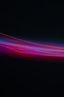 A photograph capturing vibrant streaks of light in various colors against a dark background. The lights form a wave-like pattern with predominant hues of red, purple, and blue.