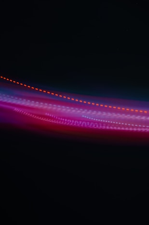 A photograph capturing vibrant streaks of light in various colors against a dark background. The lights form a wave-like pattern with predominant hues of red, purple, and blue.