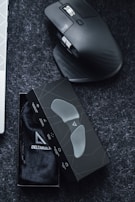 A sleek ergonomic mouse with a dark, contoured design rests on a dark surface. Next to it is a black box containing a soft pouch, possibly for the mouse or related accessories. The box features a minimalistic geometric pattern.