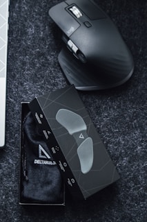 A sleek ergonomic mouse with a dark, contoured design rests on a dark surface. Next to it is a black box containing a soft pouch, possibly for the mouse or related accessories. The box features a minimalistic geometric pattern.