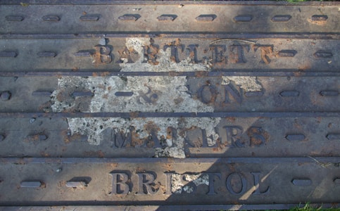 A weathered metal plate with embossed lettering, partially obscured and rusted, reads 'BARTLETT & SON MAKERS BRISTOL'. The surface shows signs of age and deterioration, with patches of rust and faded paint.