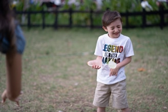 A young boy is outdoors, looking joyous as he interacts with someone off-camera. He is wearing a white T-shirt with colorful lettering saying 'Legend In The Making' and beige shorts. In his hand, he appears to be holding a small object, possibly a ball. The background features green grass and a dark fence, suggesting a park or garden setting.