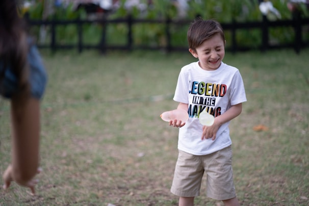 A young boy is outdoors, looking joyous as he interacts with someone off-camera. He is wearing a white T-shirt with colorful lettering saying 'Legend In The Making' and beige shorts. In his hand, he appears to be holding a small object, possibly a ball. The background features green grass and a dark fence, suggesting a park or garden setting.