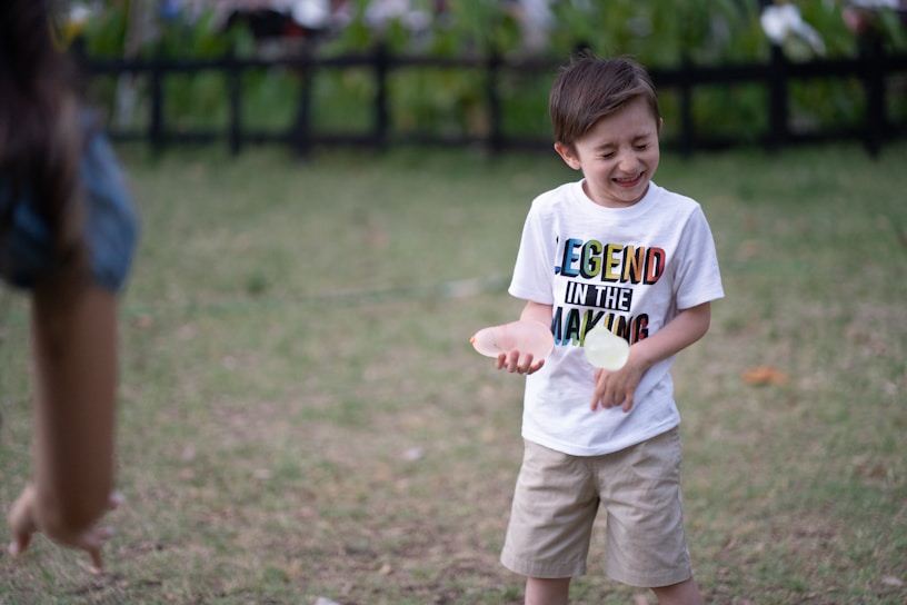 A young boy is outdoors, looking joyous as he interacts with someone off-camera. He is wearing a white T-shirt with colorful lettering saying 'Legend In The Making' and beige shorts. In his hand, he appears to be holding a small object, possibly a ball. The background features green grass and a dark fence, suggesting a park or garden setting.