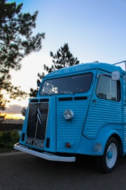A vintage blue ice cream truck parked on a road, surrounded by trees under a clear sky during sunset. The truck features classic styling with a distinct front grille and large round headlights.