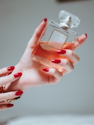 A hand with painted red nails holds a square-shaped bottle of perfume. The bottle is clear, showcasing an amber-colored liquid inside, and it has a refined, transparent cap. The setting is softly lit, creating a warm and luxurious ambiance.