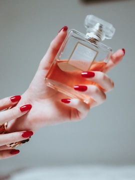 A hand with painted red nails holds a square-shaped bottle of perfume. The bottle is clear, showcasing an amber-colored liquid inside, and it has a refined, transparent cap. The setting is softly lit, creating a warm and luxurious ambiance.