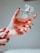 A hand with painted red nails holds a square-shaped bottle of perfume. The bottle is clear, showcasing an amber-colored liquid inside, and it has a refined, transparent cap. The setting is softly lit, creating a warm and luxurious ambiance.