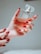 A hand with painted red nails holds a square-shaped bottle of perfume. The bottle is clear, showcasing an amber-colored liquid inside, and it has a refined, transparent cap. The setting is softly lit, creating a warm and luxurious ambiance.