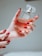 A hand with painted red nails holds a square-shaped bottle of perfume. The bottle is clear, showcasing an amber-colored liquid inside, and it has a refined, transparent cap. The setting is softly lit, creating a warm and luxurious ambiance.
