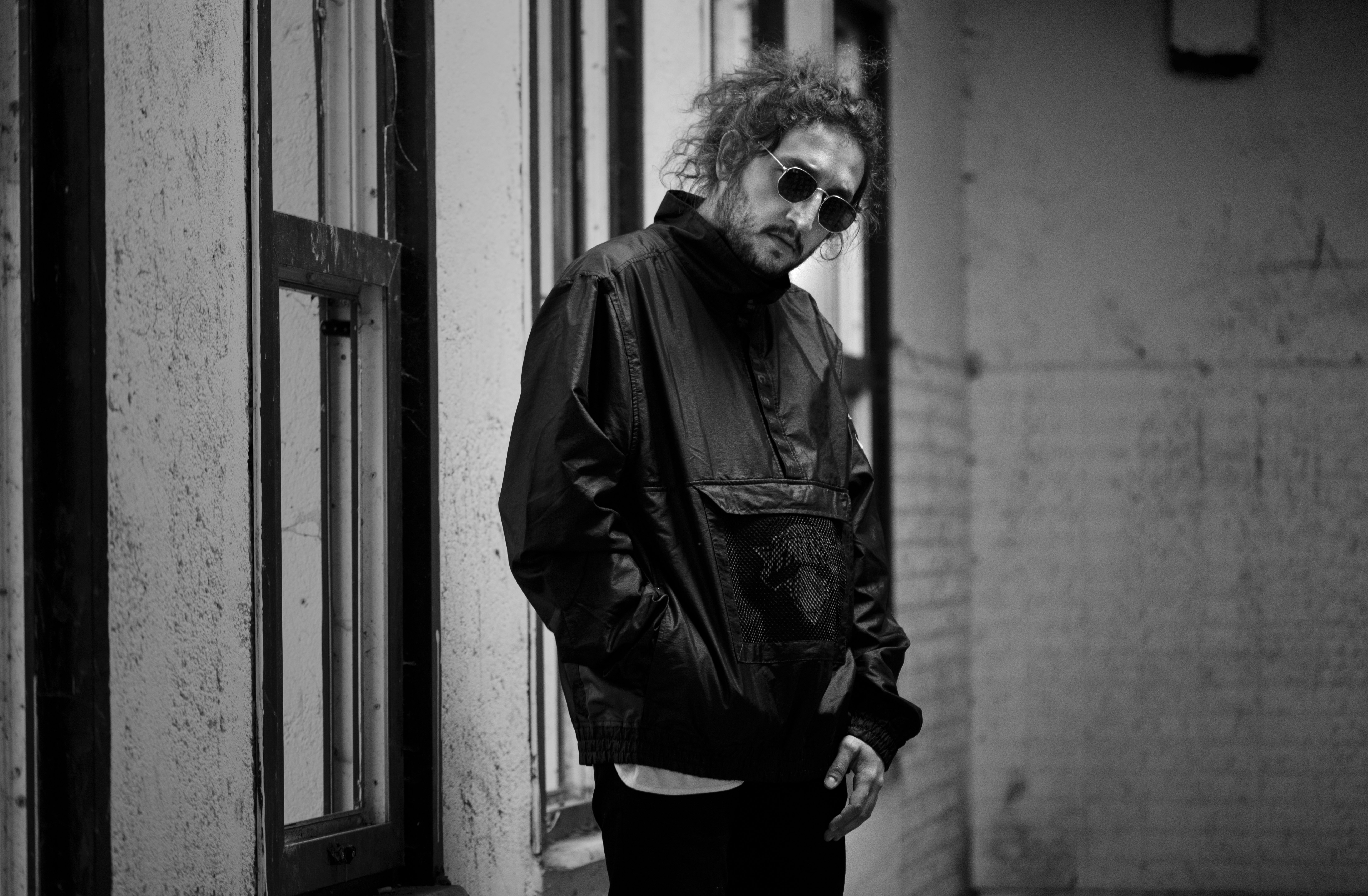 grayscale photo of woman in jacket and sunglasses standing beside wall