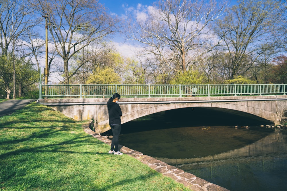 Mann in schwarzer Jacke und schwarzer Hose, der tagsüber auf einem grünen Rasenfeld in der Nähe der Brücke steht