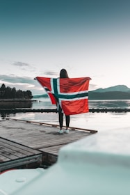 A person stands on a wooden dock near the water, holding a large Norwegian flag. The backdrop includes a serene landscape with a calm body of water and distant mountains under a clear sky.