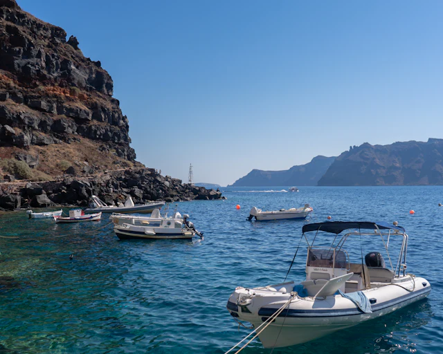 Small fishing and tour boats anchored in the clear turquoise cove of Ammoudi Bay beneath the red volcanic cliffs.