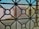 A view through a window screen featuring a geometric iron grille design. The pattern consists of intersecting circles and straight lines, creating a grid-like structure. In the background, there are blurred shapes of buildings with muted colors such as beige and light blue, suggesting an urban or residential setting.