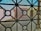 A view through a window screen featuring a geometric iron grille design. The pattern consists of intersecting circles and straight lines, creating a grid-like structure. In the background, there are blurred shapes of buildings with muted colors such as beige and light blue, suggesting an urban or residential setting.