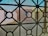 A view through a window screen featuring a geometric iron grille design. The pattern consists of intersecting circles and straight lines, creating a grid-like structure. In the background, there are blurred shapes of buildings with muted colors such as beige and light blue, suggesting an urban or residential setting.