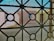 A view through a window screen featuring a geometric iron grille design. The pattern consists of intersecting circles and straight lines, creating a grid-like structure. In the background, there are blurred shapes of buildings with muted colors such as beige and light blue, suggesting an urban or residential setting.