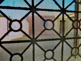 A view through a window screen featuring a geometric iron grille design. The pattern consists of intersecting circles and straight lines, creating a grid-like structure. In the background, there are blurred shapes of buildings with muted colors such as beige and light blue, suggesting an urban or residential setting.