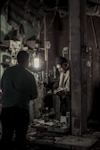 Inside a dimly lit workshop, two individuals are engaged in a task. Shelves on the walls are filled with various small boxes and tools, and a light illuminates the workspace. A man in a knit cap is handling something in his hands, standing near another person, possibly engaged in a conversation or transaction.