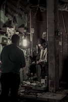 Inside a dimly lit workshop, two individuals are engaged in a task. Shelves on the walls are filled with various small boxes and tools, and a light illuminates the workspace. A man in a knit cap is handling something in his hands, standing near another person, possibly engaged in a conversation or transaction.