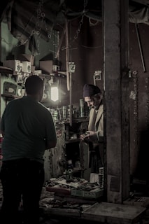 Inside a dimly lit workshop, two individuals are engaged in a task. Shelves on the walls are filled with various small boxes and tools, and a light illuminates the workspace. A man in a knit cap is handling something in his hands, standing near another person, possibly engaged in a conversation or transaction.