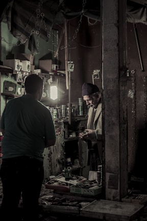 Inside a dimly lit workshop, two individuals are engaged in a task. Shelves on the walls are filled with various small boxes and tools, and a light illuminates the workspace. A man in a knit cap is handling something in his hands, standing near another person, possibly engaged in a conversation or transaction.