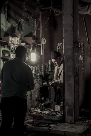 Inside a dimly lit workshop, two individuals are engaged in a task. Shelves on the walls are filled with various small boxes and tools, and a light illuminates the workspace. A man in a knit cap is handling something in his hands, standing near another person, possibly engaged in a conversation or transaction.