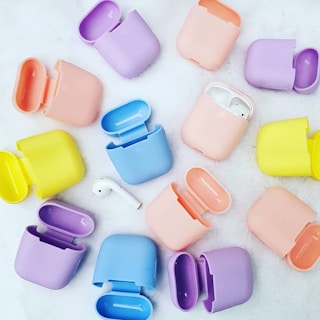 A collection of pastel-colored silicone cases for wireless earbuds are scattered across a white surface. Each case is open, showing a range of colors including lavender, pink, yellow, blue, and peach. A single earbud is visible among the cases.