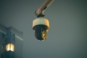 A surveillance camera is mounted on an extended arm against a background of a building at night. The building has a few lit windows, and the overall scene is dimly lit.