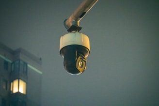 A surveillance camera is mounted on an extended arm against a background of a building at night. The building has a few lit windows, and the overall scene is dimly lit.